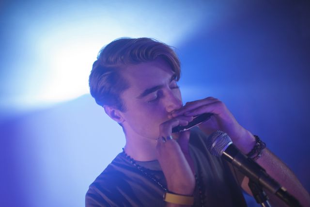 Young male musician playing harmonica on stage in a nightclub with blue lighting. Ideal for use in articles, blogs, and promotional materials about live music, nightlife, young artists, and musical performances.