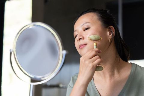 Mature african american woman using jade roller at vanity mirror for daily skincare routine