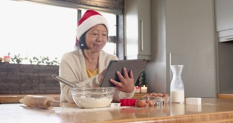 Senior Woman with Santa Hat Baking Using Tablet in Holiday Kitchen