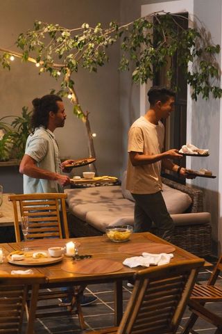 Friends carrying plates on lit patio for casual alfresco dinner party under string lights