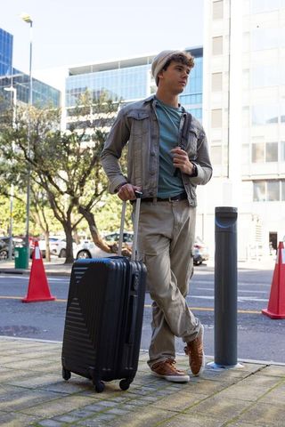 Young traveler standing on urban curb with wheeled luggage and casual street style