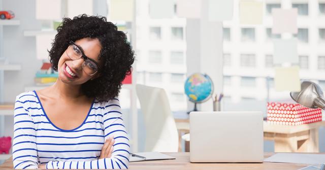 This image depicts a cheerful woman with curly hair and spectacles, smiling while sitting at a school desk. The background includes educational elements such as a globe, books, and a laptop, suggesting a classroom or academic environment. This image is ideal for use in educational materials, school websites, teacher profiles, and promotional content for academic institutions.