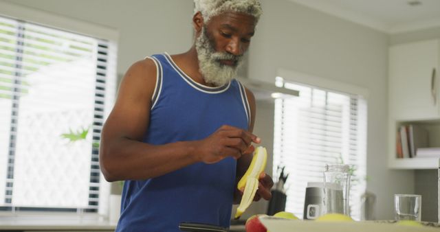 Senior man wearing a blue tank top peeling a banana in a modern, brightly lit kitchen. He appears to be preparing a healthy snack or breakfast. Perfect for uses related to healthy lifestyle, senior health, home life, nutrition, and food preparation.