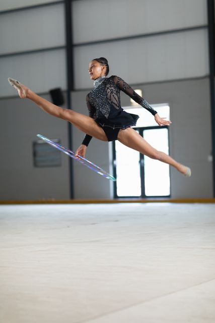 Biracial female gymnast practicing at the gym, jumping and doing splits in the air with a hula hoop in her hand. Gymnast training hard for competition.