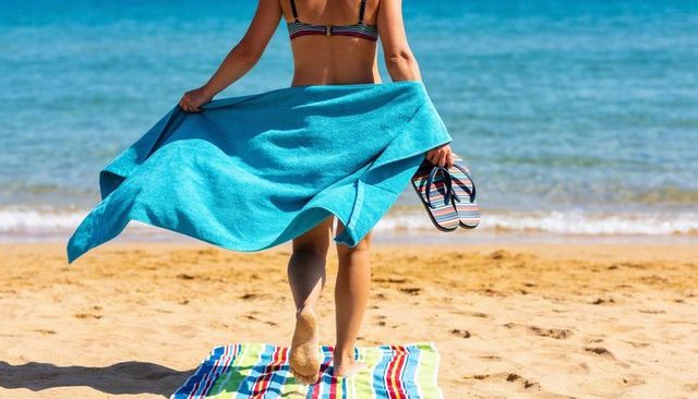 Woman walking toward ocean holding turquoise towel and striped flip-flops on sandy beach