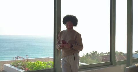 Mid-30s man standing with smartphone in modern coastal condo overlooking ocean view