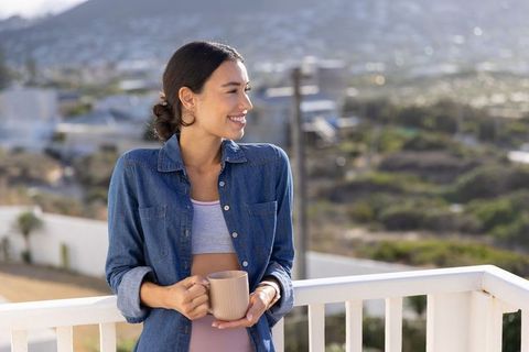 Smiling woman holding coffee mug overlooking coastal hills from balcony in denim shirt