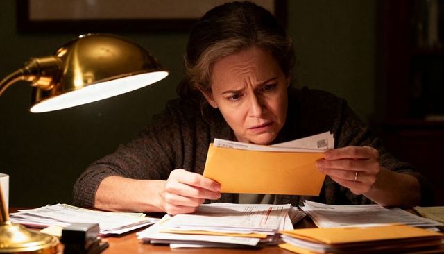 Middle-aged woman reading bill envelope at desk under brass lamp, worried about finances