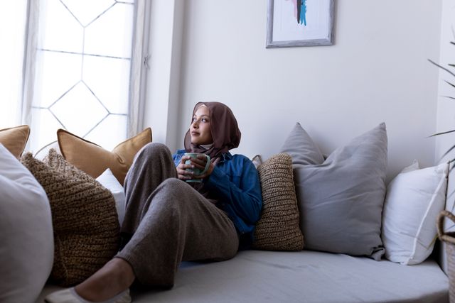 Happy biracial woman in hijab holding coffee, sitting on couch in living room at home, copy space. Happiness, health, relaxation, inclusivity and domestic life.
