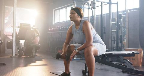 Mid adult woman resting between dumbbell sets on bench in sunlit gym with haze and focus