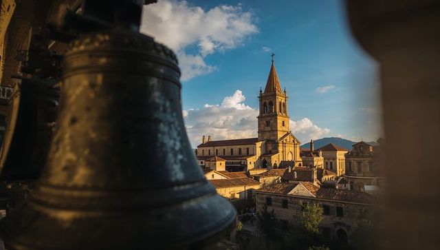 Sunlit cathedral bell tower framed by weathered bronze bell over historic tiled rooftops