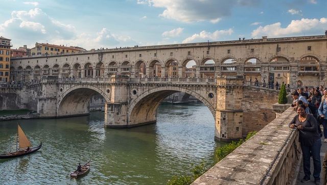 Historic arched stone bridge spanning river, sailboats gliding, pedestrians strolling promenade