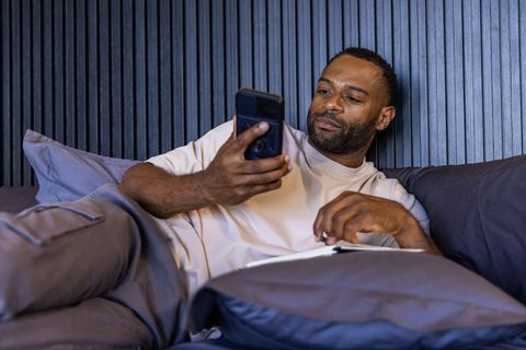 Mature man relaxing in bed using smartphone and writing in journal during cozy morning