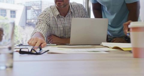 Mature man reaching for documents during collaborative office meeting with laptop and coffee