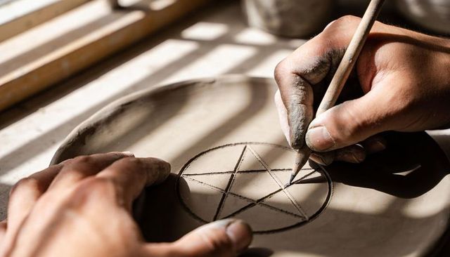 Crafting pentagram on clay plate pottery workbench, close-up hands etching ceramic