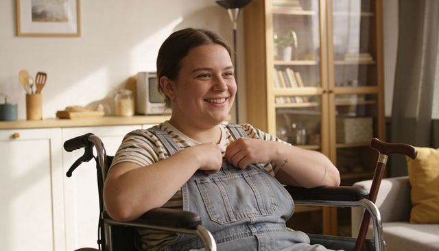 Young woman in wheelchair smiling, adjusting overalls straps, cane nearby in cozy sunlit home