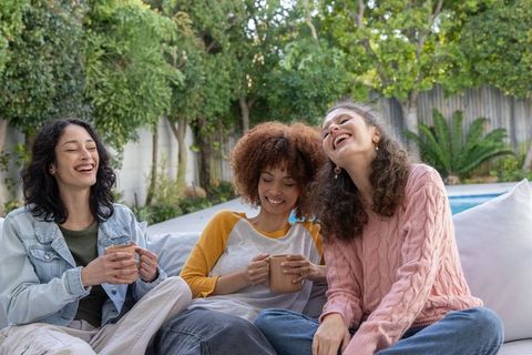 Multicultural friends laughing and relaxing on sunny poolside sofa sharing coffee and chat