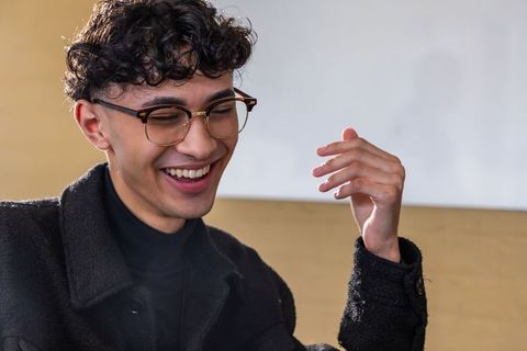 Young man smiling and laughing with tortoise glasses and raised hand, indoor portrait