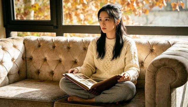 Sunlit autumn moment: woman sitting on tufted velvet sofa holding memory album reminiscing