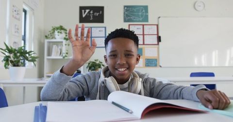Smiling schoolboy raising hand while studying with headphones and open notebook