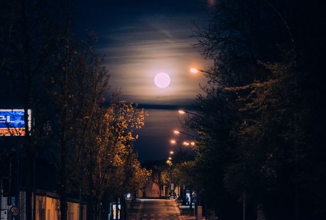 Full moon glowing brightly over a quiet city street at night. Streetlights create an ambient glow, illuminating the road with warm light. Trees outline the road, adding to the serene atmosphere. This image is suitable for themes about urban life, tranquility, and the beauty of night-time environments. Perfect for use in blogs, articles, wallpapers, and social media content related to nightly scenery, quiet moments, and serene urban settings.