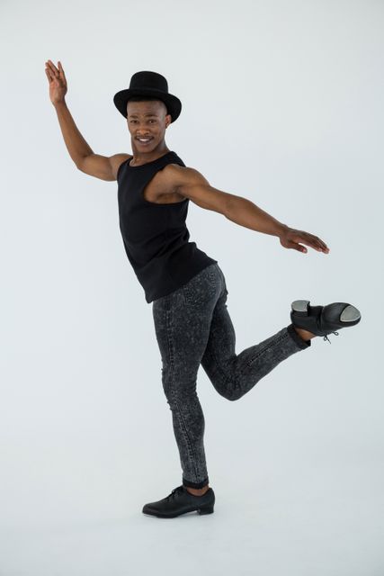 Male dancer practising dance moves in a studio, wearing a black hat, black tank top, grey pants, and tap shoes. Ideal for use in articles about dance, performance arts, dance training, and artistic expression. Suitable for promoting dance classes, workshops, and dance-related events.