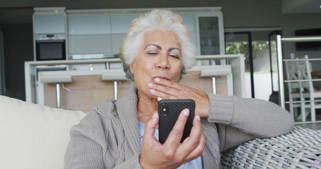 Senior woman with grey hair making a video call on a smartphone while sitting on a comfortable couch in a modern home. She appears engaged and connected, touching her cheek while looking at the phone. Ideal for use in advertisements or articles about elderly people using technology and staying connected with loved ones or for promoting video calling services or mobile phone usage among senior citizens.