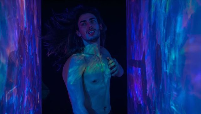Turning shirtless male posing under neon projection lights casting iridescent blue-purple glow