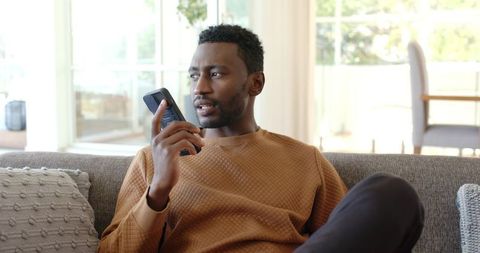 African American man speaking into cordless phone while relaxing on gray living room sofa