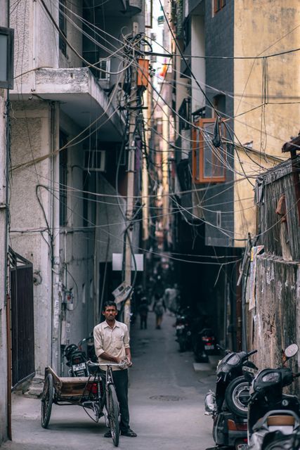A man is standing with his bicycle rickshaw in a narrow alleyway in an Indian urban area. Visible wires and scooters indicate an older, bustling neighborhood. This scene is ideal for depicting daily life in India, showcasing traditional transportation, urban living, and local culture in Southeast Asia. It can be used in articles and projects related to travel, community life, and urban infrastructure.