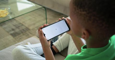 African american boy holding smartphone on sofa at home with blank screen for app mockup, mobile gam
