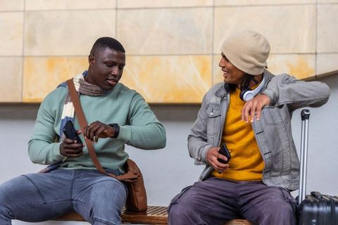 Two men waiting at transit bench checking watch and phones with luggage and headphones