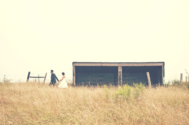 Couple holding hands and walking through a grassy field near a wooden shelter, evoking feelings of romance and togetherness in a rural setting. Suitable for depicting romantic themes, relationships, or advertising rustic getaways.