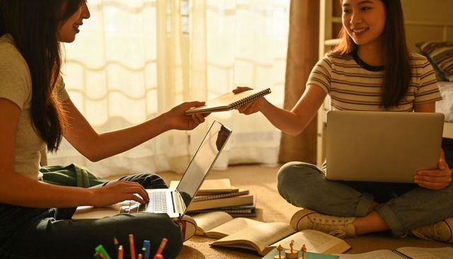Teen girls studying together in cozy dorm room exchanging notebook while using laptops