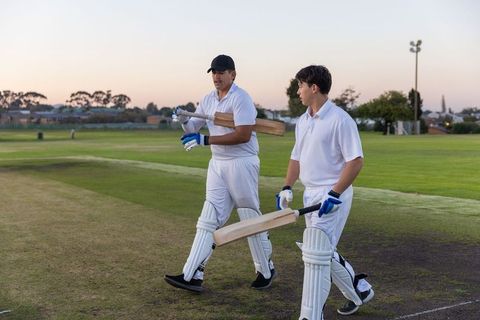 Cricket players walking off field at dusk carrying bats and pads after match