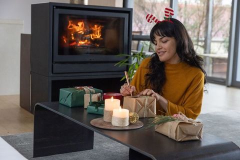 Woman wrapping christmas gifts by fireplace with candles and decorations