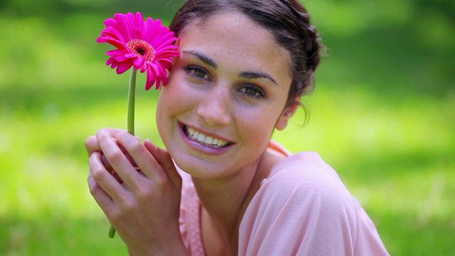 A cheerful young woman with a pink flower, outside in a sunny park. This can be used for advertisements promoting outdoor activities, nature walks, or beauty products. Perfect for blogs and articles about happiness, lifestyle, and well-being.