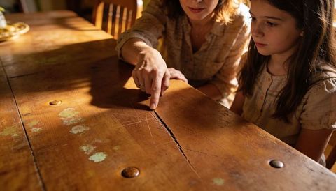 Mother pointing at crack on rustic wooden table while daughter observes in sunlight