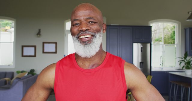 Charming elderly man with grey beard wearing red shirt is smiling warmly in contemporary home interior. Ideal for illustrating themes of active aging, happiness, domestic life, and fitness. Perfect for advertisements, articles, and marketing materials emphasizing positive attitudes and active lifestyles among seniors.