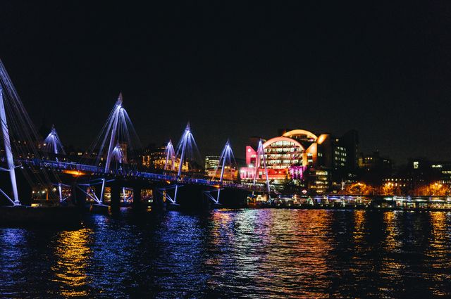 This stock photo showcases a vibrant cityscape captured at night with an illuminated bridge creating impressive reflections on the river. Charismatic city lights complement the urban atmosphere, emphasizing modern architecture. Ideal for websites or promotions related to city tourism, urban nightlife, architecture highlights, or event promotions highlighting nocturnal city decorating.