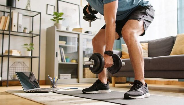 Man squatting with adjustable dumbbells in living room, home workout with laptop