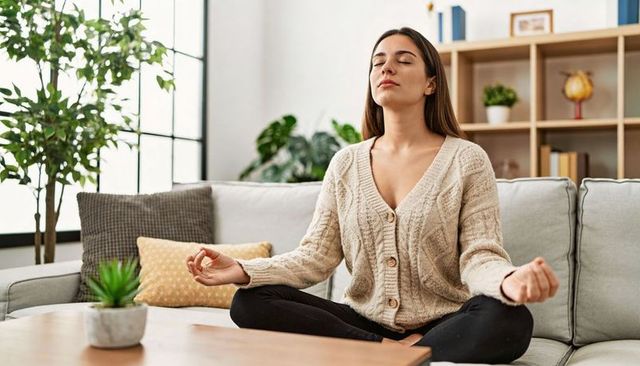 Young woman meditating cross-legged on cozy sofa in sunlit neutral living room