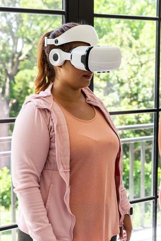 African american woman wearing vr headset and headphones near glass doors in pink jacket