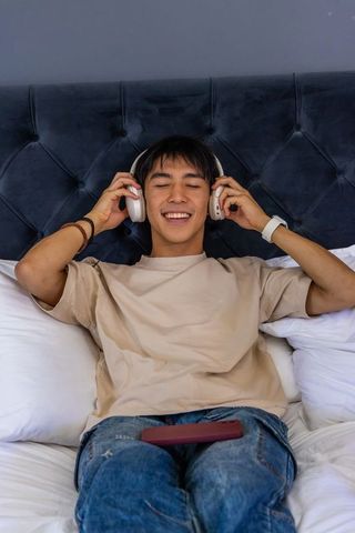 Young asian man relaxing on bed listening to music with wireless headphones and smartphone