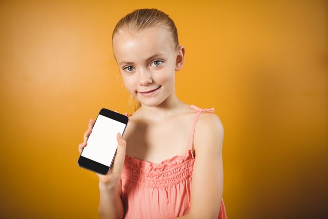 Young girl smiling and holding a smartphone with a blank screen against an orange background. Ideal for technology, communication, and lifestyle themes. Perfect for advertisements, educational materials, and social media content.