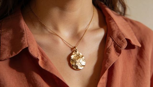 Featuring hammered gold pendant necklace on woman collarbone wearing rust shirt in warm light