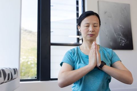 Asian woman practicing mindfulness meditation at home wearing earbuds and smartwatch