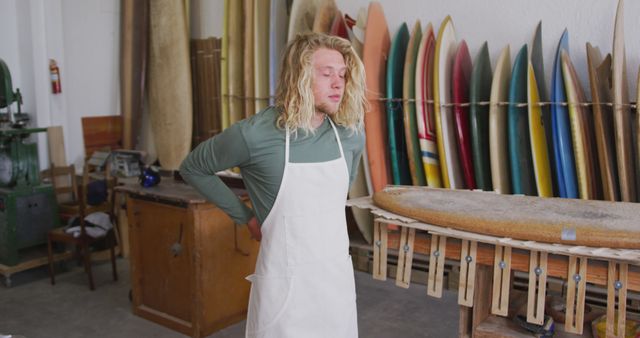 Caucasian male surfboard maker with long blonde hair putting on apron at workshop, unaltered. Small business, work, sports equipment and craftsmanship.