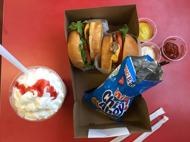 Fast food meal includes two burgers with lettuce, cheese, tomato, three sauce cups, a bag of cookies, and a milkshake with whipped cream and cherry syrup on top. Perfect for illustrating takeout, lunchtime, or snack options. Can be used for restaurant menus, food delivery services, or promotions for fast food chains.