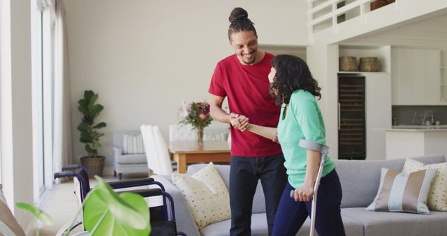 Smiling biracial man with dreadlocks helping female partner to wheelchair in living room. wellbeing and domestic lifestyle with physical disability.